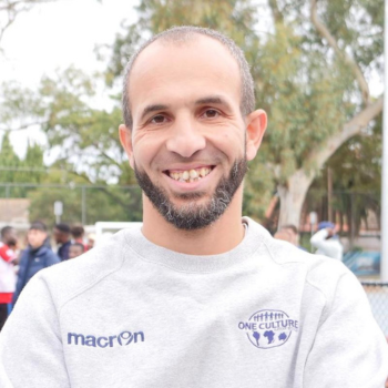 Portrait of a smiling man with a beard wearing a grey One Culture sweatshirt outdoors.